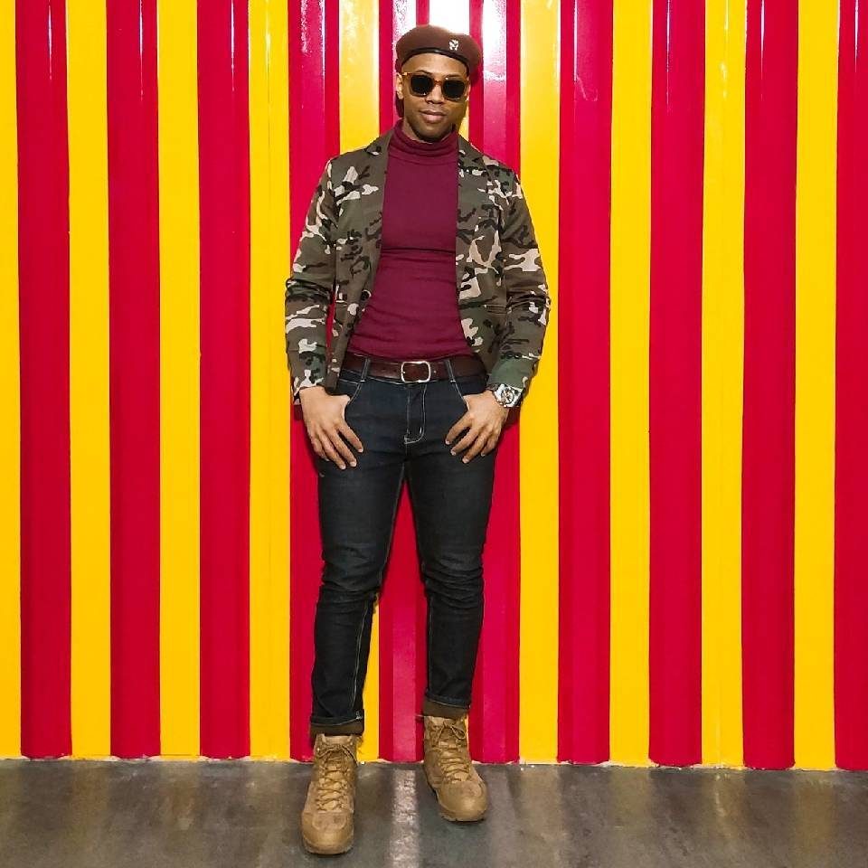 Danny Flight in a beret, sunglasses, and camo blazer standing against a red and yellow striped wall.
